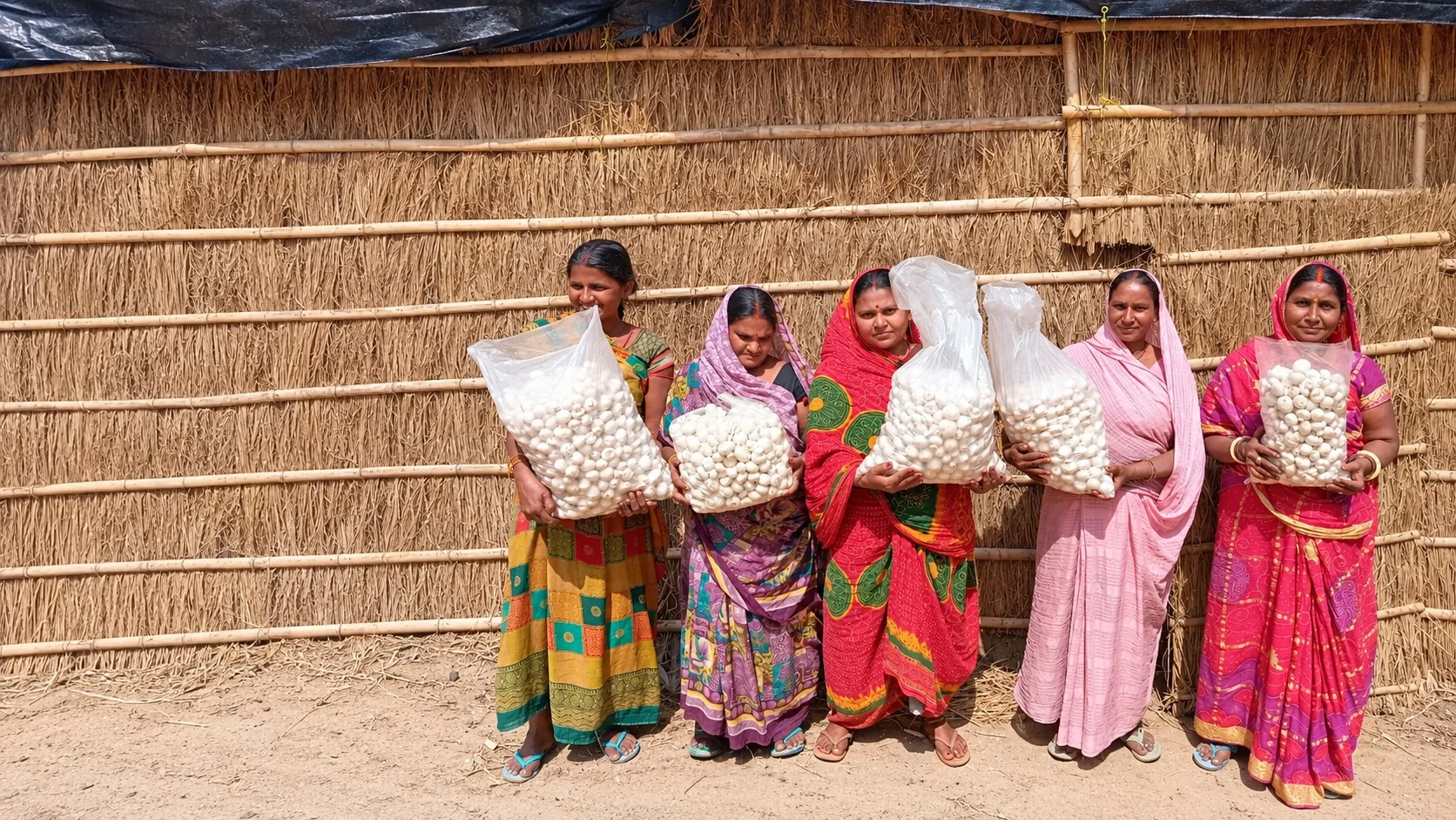 Five community mushroom farmers holding their harvest outside a bamboo and grass hut
